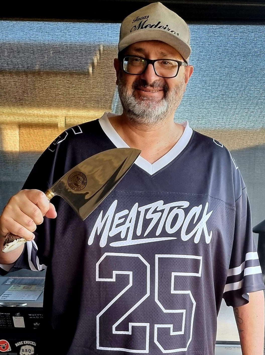 Man wearing a 'Meatstock 25' jersey holding a MEDEIROS CLEAVER in front of a barbecue grill.