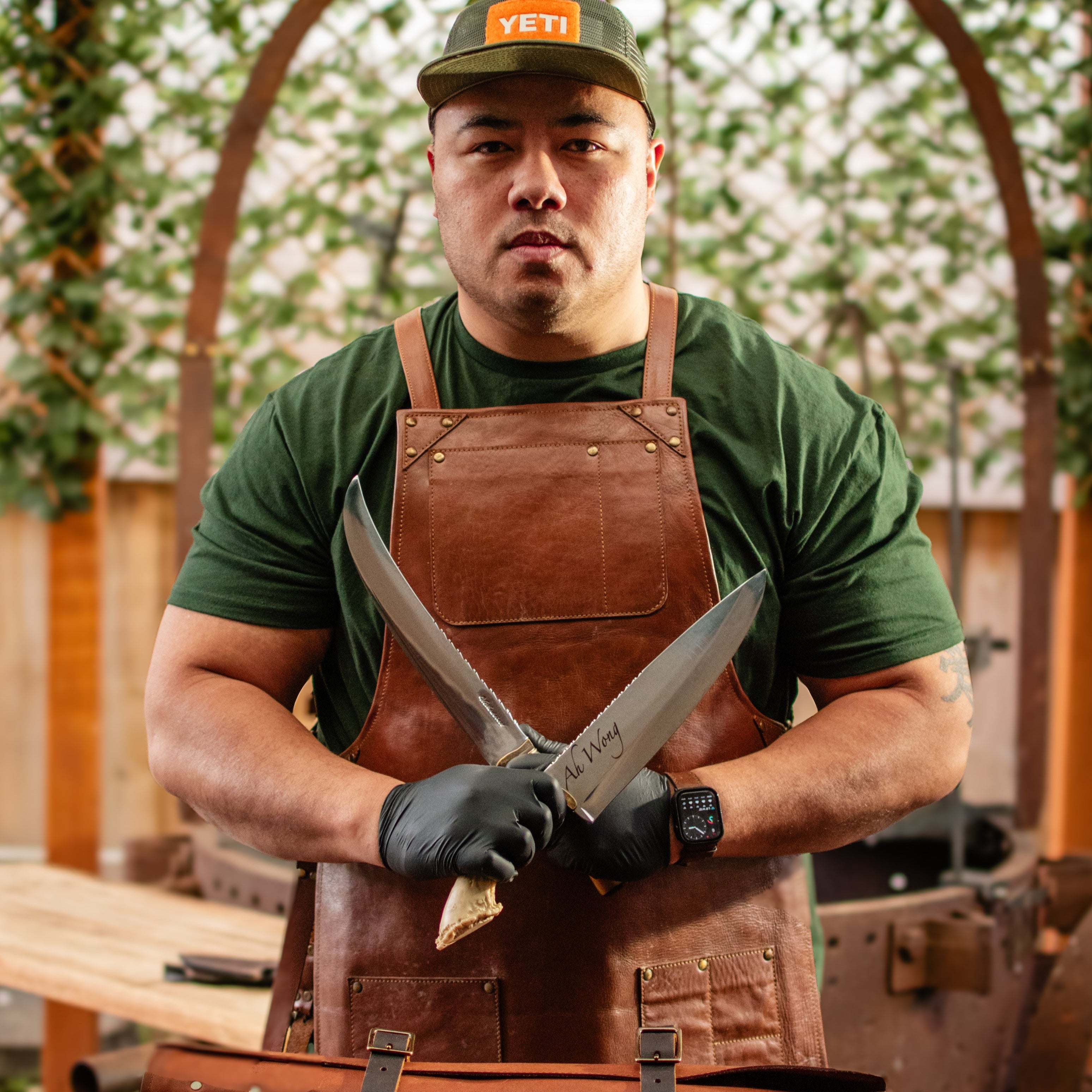 Man wearing a green shirt, brown apron, and YETI cap holding two large Medeiros knives outdoors.